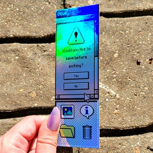 Pode incluir: Um marcador de livros retangular com um design de interface de computador retro. O marcador de livros exibe uma caixa de di&aacute;logo com o texto "Would you like to save before exiting?" e inclui &iacute;cones para uma imagem, uma pasta, informa&ccedil;&otilde;es e uma lixeira.