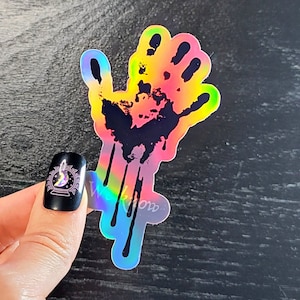 May include: A holographic sticker of a black handprint with dripping blood. The handprint is outlined in rainbow colors.