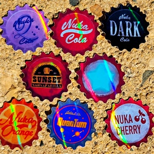 May include: A collection of seven bottle caps with various designs and text. The caps feature the names "Nuka Cola", "Nuka Dark Cola", "Nuka Orange", "Nuka Cherry", "Nuka Quantum", and "Sunset Sarsaparilla". Each cap has a unique color scheme.