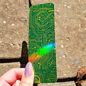 May include: A green and yellow circuit board bookmark. The bookmark has a complex pattern of lines and shapes, resembling the internal components of a computer. The bookmark is held against a textured, gray surface.