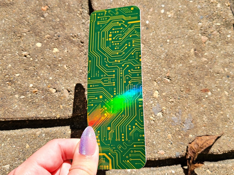 May include: A green and yellow circuit board bookmark. The bookmark has a complex pattern of lines and shapes, resembling the internal components of a computer. The bookmark is held against a textured, gray surface.