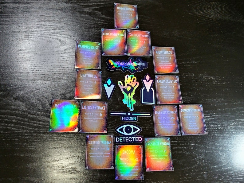May include: A collection of holographic cards with various names and descriptions. The cards are arranged in a triangular shape on a wooden surface. The cards feature text such as "Tundra Cotton", "Luna Moth Wing", "Vampire Dust", "Deathbell", "Moon Sugar", "Lotus Extract", "Sleeping Tree Sap", "Skooma", "Torchbug Thorax", "Nightshade", "Creep Cluster", "Dragon's Tongue", "Soul Husk Extract", "Frostbite Venom", and "Watch the skies, Traveler."