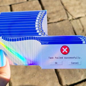 May include: A stack of iridescent stickers featuring a blue and white Windows error message. The message reads "Error" and "Task failed successfully." with a red X icon. The buttons "OK" and "Cancel" are visible.