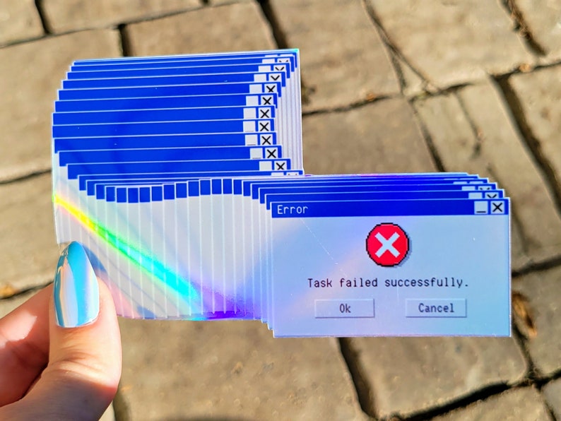 May include: A stack of iridescent stickers featuring a blue and white Windows error message. The message reads "Error" and "Task failed successfully." with a red X icon. The buttons "OK" and "Cancel" are visible.