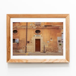 May include: A framed photograph of a building facade with a brown door and an anchor. The building has a weathered, orange-toned exterior with two circular windows. A sign above the door is visible. The photograph is in a light-colored wooden frame.