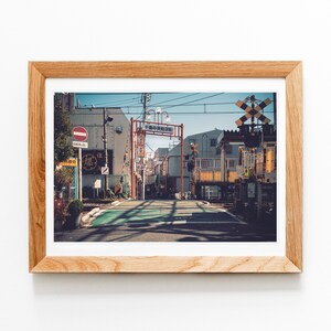 May include: A framed print of a Japanese street scene. The image features a road with a green crosswalk, buildings, and a railway crossing. The sky is blue, and the frame is made of light-colored wood.