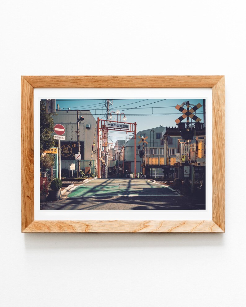 May include: A framed print of a Japanese street scene. The image features a road with a green crosswalk, buildings, and a railway crossing. The sky is blue, and the frame is made of light-colored wood.