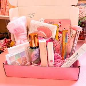 以下が含まれることがあります： ピンクの段ボール箱に入った美容製品セット。ピンクのぬいぐるみヘッドバンド、マルチカラーのコンシーラーパレット、ポンプボトル、Sweet Mintの箱、ビューラー、キラキラしたゴールドのチューブ、ピンクのジェイドローラー、4チップアイブロウペンが含まれています。