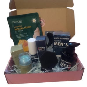 Op de afbeelding: Een cadeaupakket met verzorgingsproducten voor mannen. Inclusief een voetmasker, gezichtscrème, gezichtsreiniger en andere huidverzorgingsartikelen. De roze doos is gevuld met papier.