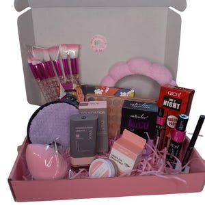 Puede incluir: Una caja de regalo rosa llena de productos de maquillaje, incluyendo brochas, base, sombra de ojos y máscara de pestañas. La caja contiene una esponja de maquillaje, un iluminador de ojos y una diadema.