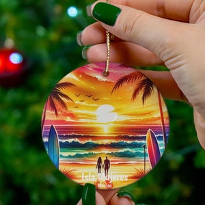 May include: A round ceramic ornament with a sunset scene over the ocean. The ornament features a couple walking on the beach with surfboards in the water. The text "Isla Mujeres, Mexico" is printed on the ornament.