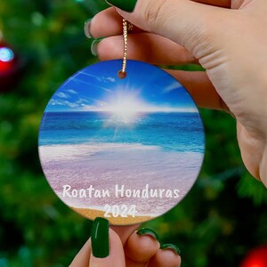 May include: A round ceramic Christmas ornament with a beach scene. The ornament features a blue sky, white clouds, a bright sun, blue ocean, and white sand. The text "Roatan Honduras 2024" is printed on the ornament.