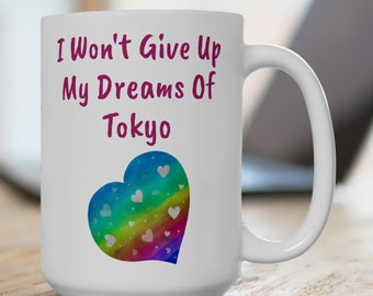 May include: White ceramic mug with a pink inscription that reads "I Won't Give Up My Dreams Of Tokyo" and a rainbow heart with white hearts inside.