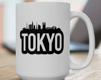 May include: White ceramic mug featuring a black silhouette of the Tokyo skyline with the city's name in bold, black letters. The mug has a large handle and is set on a wooden surface.
