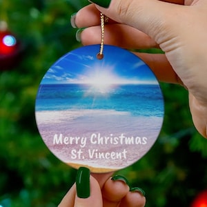 May include: A round Christmas ornament featuring a beach scene with a bright sun, blue sky, and ocean. The ornament has the text "Merry Christmas St. Vincent" in white. A gold string is attached for hanging.