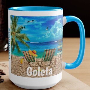 May include: White ceramic coffee mug with a blue rim and handle. The mug features a colorful image of a beach scene with two lounge chairs, a palm tree, and the text "Goleta, California".