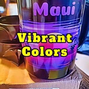 May include: A black ceramic mug with a sunset scene and the word "Maui" in purple lettering. The mug has vibrant colors of pink, purple, and blue. The text "Vibrant Colors" is superimposed on the image.