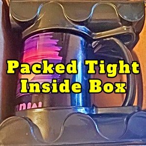 May include: A black travel mug with a purple and pink abstract design is packed tightly inside a brown cardboard box. The text "Packed Tight Inside Box" is written in yellow letters on the mug.