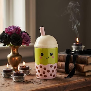 May include: A whimsical ceramic boba tea cup with a smiling face, green body, pink polka dots, and a cream-colored lid. The cup has a pink straw. The scene includes candles, books, and flowers.