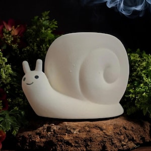 May include: White ceramic snail incense burner with a smiling face. The snail is sitting on a brown surface with green foliage in the background.