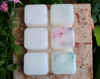 May include: A set of six wax melts in a square shape, each with a different color and glitter. The wax melts are arranged in a grid pattern on a brown surface.