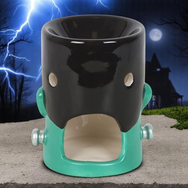 May include: A ceramic Frankenstein-themed oil warmer. The top is black, with a green face and body. It has two small holes for eyes and a rectangular opening at the bottom. The warmer has a cylindrical shape and is designed for Halloween.