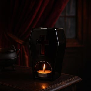 May include: A black ceramic wax warmer shaped like a coffin with a cutout cross design on the front. The base features an arched opening containing a lit tea light candle, which illuminates the interior and casts a warm glow through the cross cutout.