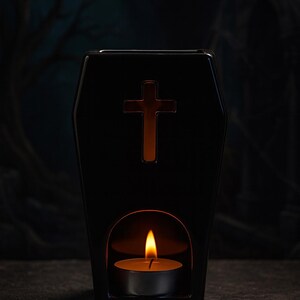 May include: A black ceramic wax warmer shaped like a coffin with a cutout cross symbol on the front. The base features an arched opening containing a lit tea light candle, which illuminates the interior and casts a warm glow through the cross.