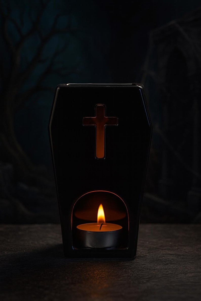 May include: A black ceramic wax warmer shaped like a coffin with a cutout cross symbol on the front. The base features an arched opening containing a lit tea light candle, which illuminates the interior and casts a warm glow through the cross.