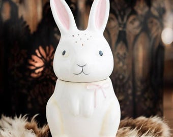 May include: White ceramic bunny figurine with pink ears and a pink bow. The bunny has a sweet expression and is sitting on a brown fur surface.