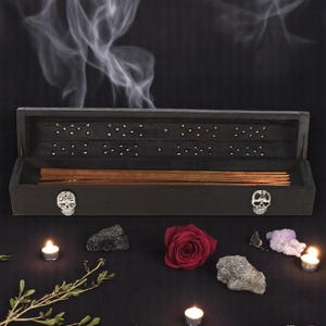 May include: A dark grey incense box with a skull motif, containing incense sticks. A red rose, crystals, and lit candles are arranged around the box. Smoke drifts upwards from the box against a black backdrop.