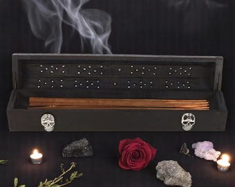 May include: A black incense box with a skull design on the front, filled with incense sticks. A red rose, crystals, and lit candles surround the box. Wisps of smoke rise from the box against a dark background.