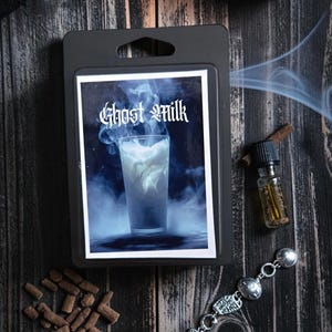 May include: A rectangular black plastic wax melt clamshell package featuring a label with the text Ghost Milk. The label displays a glass of white liquid surrounded by swirling blue smoke and mist against a dark background.