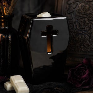 May include: A glossy black ceramic wax warmer shaped like a coffin, featuring a cutout cross design on the front. The top of the warmer holds a white wax melt, and the interior emits a warm orange glow through the cross-shaped opening.