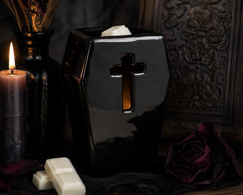 May include: A glossy black ceramic wax warmer shaped like a coffin, featuring a cutout cross design on the front. The top of the warmer holds a white wax melt, and the interior emits a warm orange glow through the cross-shaped opening.