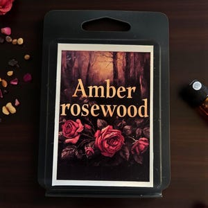 May include: A black wax melt package with a label featuring the words "Amber rosewood" in a serif font. The label also displays a dark forest scene and three red roses. A small bottle is visible in the background.