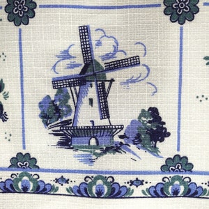 May include: A white fabric with a blue and green windmill pattern. The windmill is surrounded by clouds and trees. The fabric has a blue and green border with a floral design.