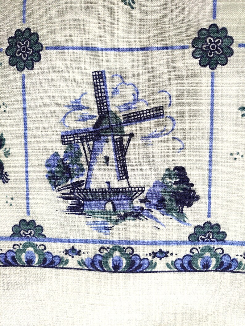 May include: A white fabric with a blue and green windmill pattern. The windmill is surrounded by clouds and trees. The fabric has a blue and green border with a floral design.