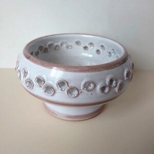 Könnte beinhalten: Weiße Keramikschale mit braunem Rand und einem dekorativen Muster aus kleinen Löchern um den oberen und unteren Rand. Die Schale steht auf einem Sockel.