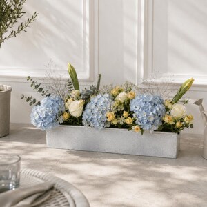 May include: A rectangular silver-toned metal planter box filled with a floral arrangement featuring light blue hydrangea clusters, cream-coloured roses, small yellow spray roses, green eucalyptus sprigs, and tall thin green stems.