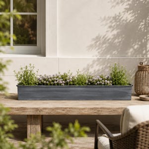 May include: A rectangular, grey metal planter box filled with green plants and small purple flowers. The planter sits on a weathered wooden table outdoors, with a window and a woven basket in the background.