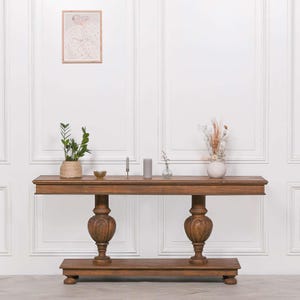 May include: A long wooden console table with a rectangular top, two large turned pedestal bases, and a bottom shelf. The table is decorated with a small potted plant, a glass bowl, two candles, a small vase, and a white vase with dried flowers.