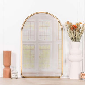 May include: An arched wall mirror with a thin gold metal frame. The mirror reflects a window with a grid pattern. To the left is a tall, ribbed, peach-coloured candle and a small glass bottle. To the right is a white vase with dried flowers.