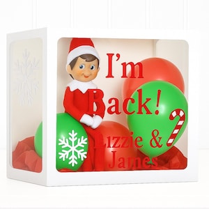 May include: A white box with a clear front panel, featuring a red and white elf doll sitting on red and green balloons. The balloons have snowflake and candy cane designs. Text on the balloons reads "I'm Back! Lizzie & James."