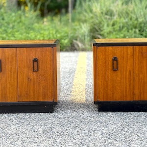 May include: Two matching wooden cabinets with a medium brown wood grain finish. Each cabinet features two doors with rectangular black metal pulls, a black top border, and a solid black base. The cabinets are positioned side by side.