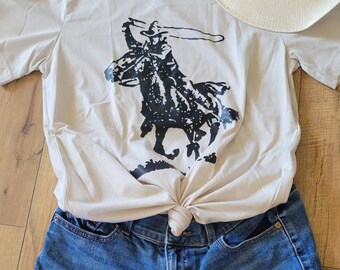 May include: A beige t-shirt with a black graphic of a cowboy roping a horse, paired with denim shorts and a straw hat. The shirt is knotted at the waist, creating a casual, stylish look. The outfit is laid out on a wooden surface.