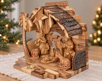 May include: A handcrafted wooden nativity scene carved from olive wood, featuring a stable structure with a bark-textured roof, a star, and figures of a family, a donkey, and a sheep. The base includes a small circular gold-toned metal inlay.