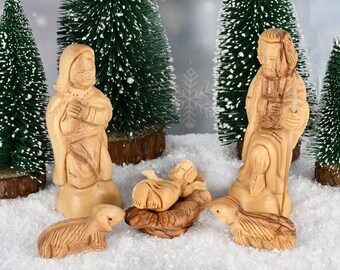 Peut inclure: Une crèche en bois avec des figurines sculptées de Marie, Joseph, l'enfant Jésus dans une mangeoire et deux agneaux. Les figurines sont placées devant des sapins de Noël miniatures et de la neige artificielle.