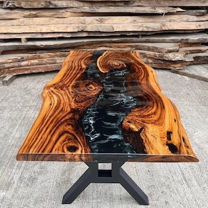 May include: A wooden table with a black epoxy resin river running through the center. The table has a live edge and a black metal base.