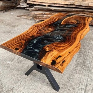 May include: A live edge wood table top with a black and white epoxy river running through the center. The table top is supported by a black metal X-shaped base.